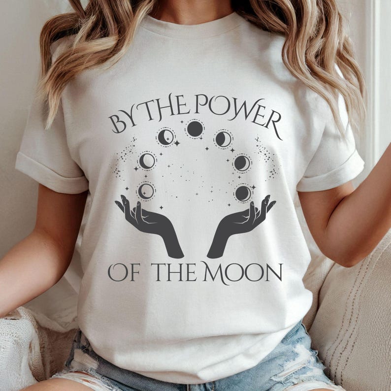 A woman wearing a white t-shirt with the text "BY THE POWER OF THE MOON" printed on it.