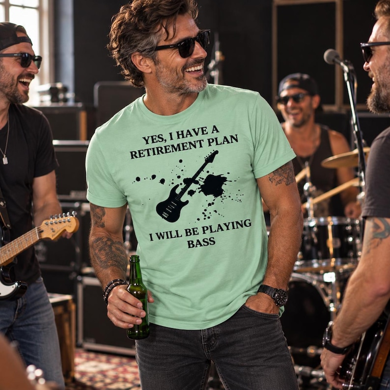 May include: A light green short-sleeved t-shirt featuring a black graphic of a bass guitar with ink splatters and the text: YES, I HAVE A RETIREMENT PLAN I WILL BE PLAYING BASS. The shirt has a crew neckline and a standard fit.