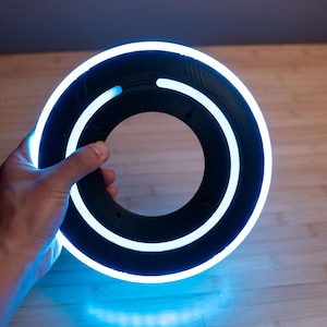 May include: A circular black disc featuring a bright, glowing blue neon ring light embedded within its structure. The object has a hollow center and a smooth, matte finish, with the light creating a vibrant, continuous circular illumination.