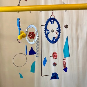 May include: A colorful mobile made of metal and plastic. It features blue and red gears, blue and green triangles, and yellow and red pom-poms. The mobile is suspended from a yellow wooden rod.