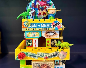 Könnte beinhalten: Ein mehrstöckiger Turm aus Pappe, der mit verschiedenen Bildern und Objekten verziert ist. Der Turm ist gelb, grün und rot und trägt ein Schild mit der Aufschrift "Deli Meat" und ein weiteres mit der Aufschrift "Salon". Der Turm ist mit verschiedenen Bildern verziert, darunter ein Dinosaurier, ein Augenpaar, ein Dollar-Schein und ein Bild von zwei Engeln. Der Turm ist mit einem gelben Pompon und einer blauen Feder gekrönt.