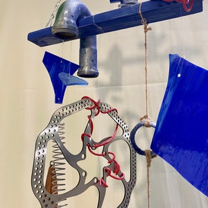 May include: A blue and silver mobile with a metal brake rotor, a blue glass piece, and a metal pipe. The mobile is suspended from a wooden beam.