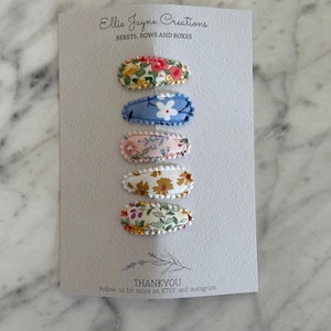 May include: Set of five floral fabric hair clips. The clips are oval shaped and have a white scalloped edge. The floral patterns are different on each clip. The clips are arranged vertically on a white card with the text "Ellie Jayne Creations" and "BERETS, BOWS AND BOXES" at the top and "THANKYOU" and "Follow us for more on ETSY and instagram" at the bottom.