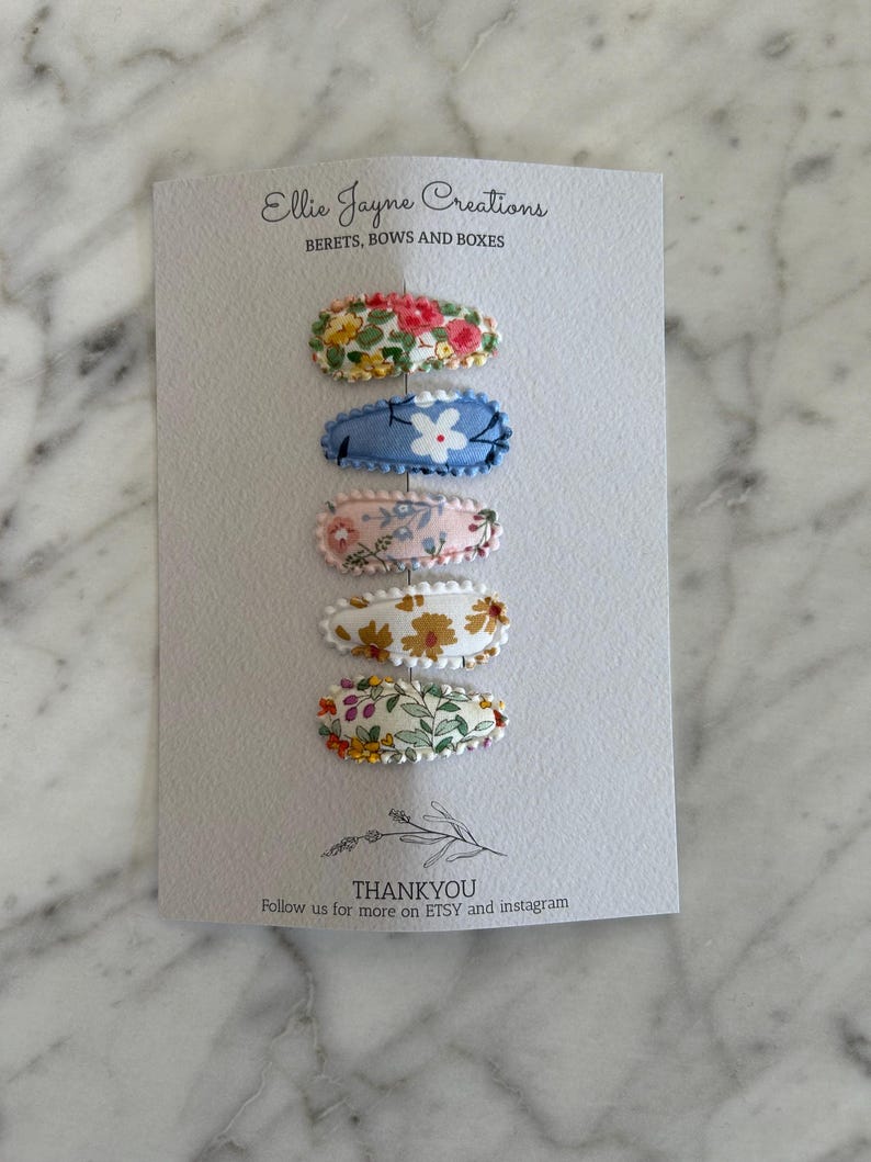 May include: Set of five floral fabric hair clips. The clips are oval shaped and have a white scalloped edge. The floral patterns are different on each clip. The clips are arranged vertically on a white card with the text "Ellie Jayne Creations" and "BERETS, BOWS AND BOXES" at the top and "THANKYOU" and "Follow us for more on ETSY and instagram" at the bottom.