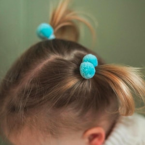 May include: A young girl with blonde hair has two blue pom-pom hair ties in her hair.