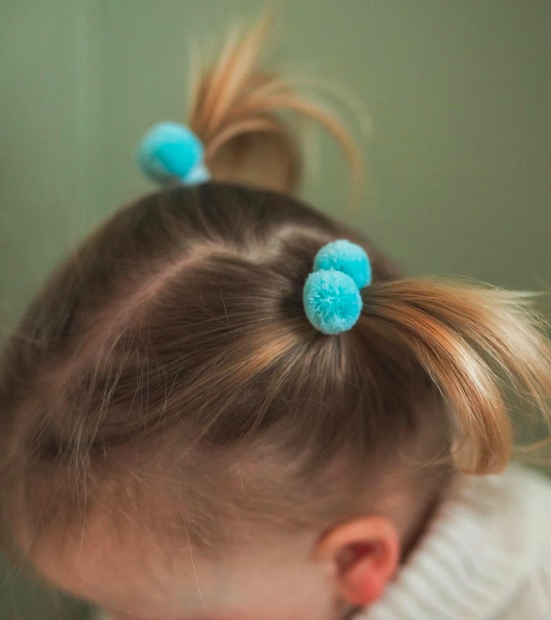 May include: A young girl with blonde hair has two blue pom-pom hair ties in her hair.