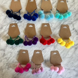 May include: Twelve sets of three pom-pom hair ties on brown card stock. The pom-poms are in various colours including blue, green, red, yellow, pink, and purple.