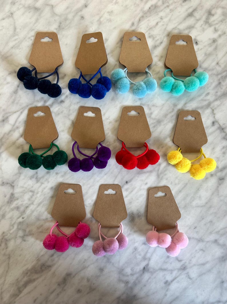 May include: Twelve sets of three pom-pom hair ties on brown card stock. The pom-poms are in various colours including blue, green, red, yellow, pink, and purple.