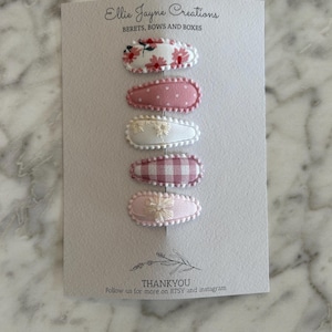 May include: Set of six pink and white fabric hair clips with floral and gingham patterns. The clips are displayed on a white card with the text "Ellie Jayne Creations" and "BERETS, BOWS AND BOXES".