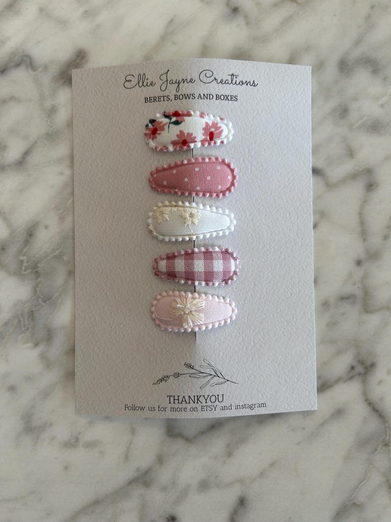 May include: Set of six pink and white fabric hair clips with floral and gingham patterns. The clips are displayed on a white card with the text "Ellie Jayne Creations" and "BERETS, BOWS AND BOXES".