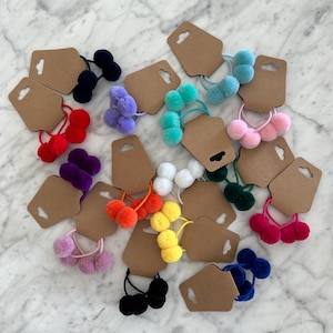 May include: Assortment of colourful fuzzy pom-pom hair ties on brown card stock tags. The hair ties are in various colours including red, blue, green, purple, pink, yellow, orange, and black.