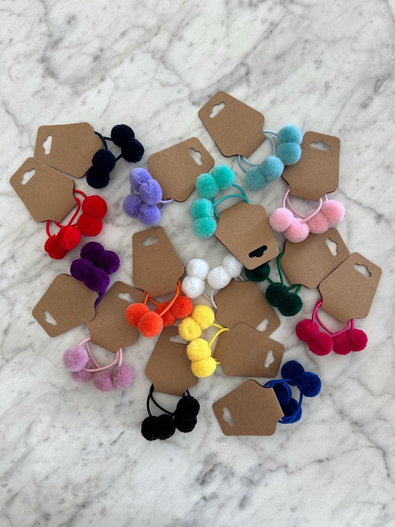 May include: Assortment of colourful fuzzy pom-pom hair ties on brown card stock tags. The hair ties are in various colours including red, blue, green, purple, pink, yellow, orange, and black.