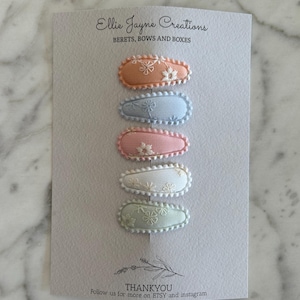 May include: Set of six pastel-colored hair clips with floral embroidery. The clips are arranged vertically, with the top clip being peach, followed by light blue, pink, white, and light green. The clips are all oval-shaped and have a scalloped edge.