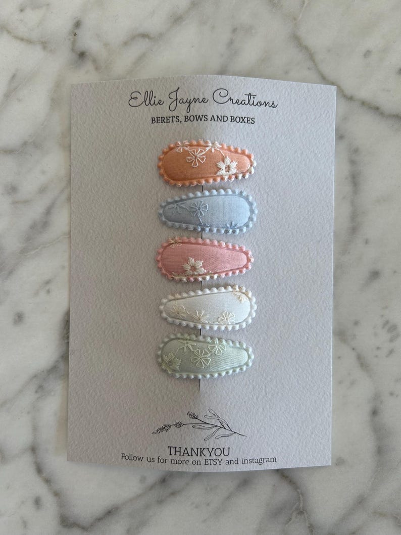 May include: Set of six pastel-colored hair clips with floral embroidery. The clips are arranged vertically, with the top clip being peach, followed by light blue, pink, white, and light green. The clips are all oval-shaped and have a scalloped edge.