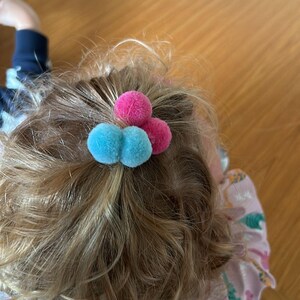 May include: A close-up of a child's hair with two blue and one pink pom-pom hair ties.