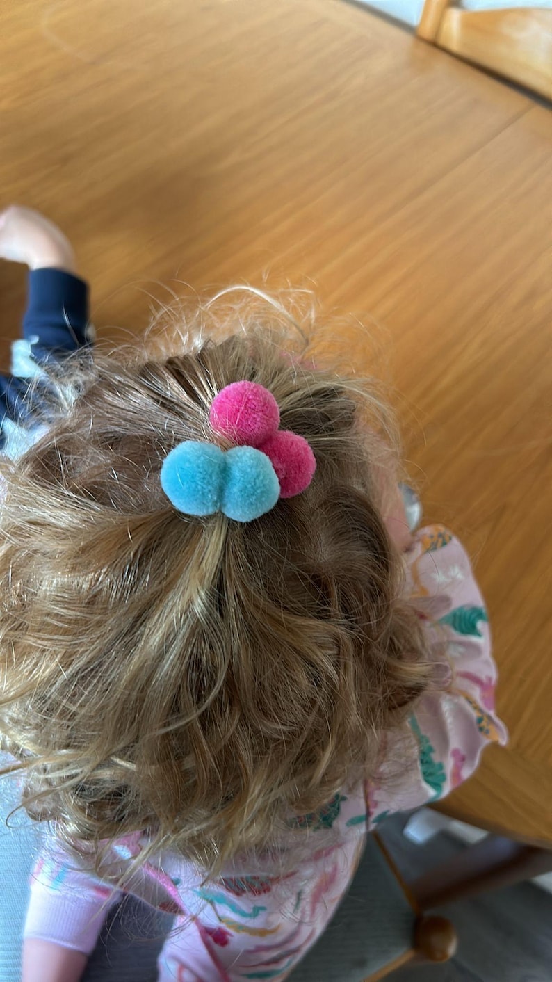 May include: A close-up of a child's hair with two blue and one pink pom-pom hair ties.