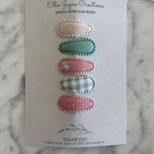 May include: Set of six fabric-covered hair clips in pink, green, and white patterns. The clips are arranged vertically on a white card with the text "Ellie Jayne Creations" and "BERETS, BOWS AND BOXES".