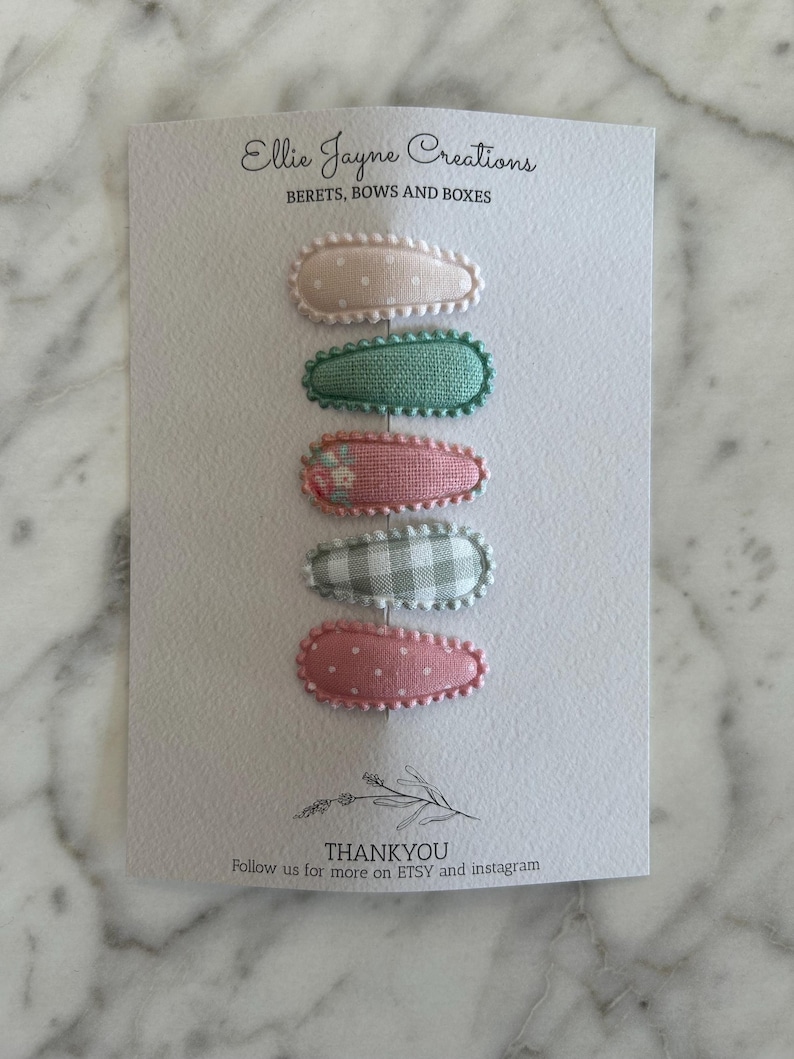 May include: Set of six fabric-covered hair clips in pink, green, and white patterns. The clips are arranged vertically on a white card with the text "Ellie Jayne Creations" and "BERETS, BOWS AND BOXES".