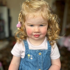 May include: A young girl with long, curly blonde hair wearing a white t-shirt and a blue denim pinafore dress with a floral embroidery design. She has a pink bow in her hair.