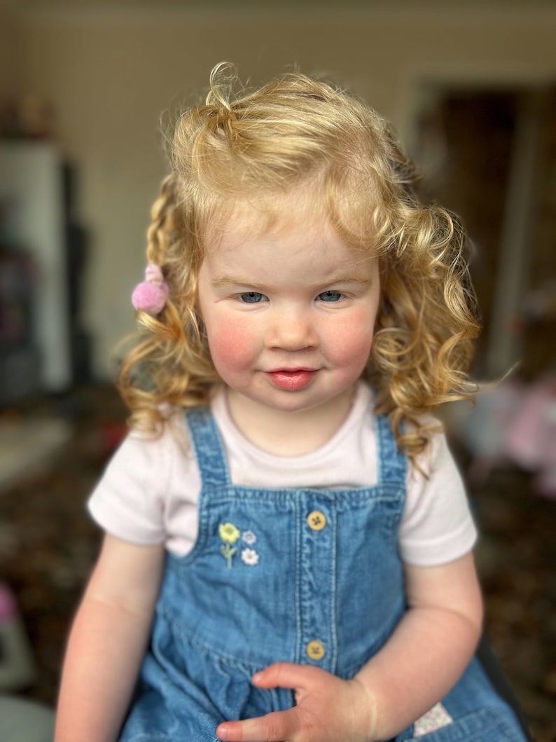 May include: A young girl with long, curly blonde hair wearing a white t-shirt and a blue denim pinafore dress with a floral embroidery design. She has a pink bow in her hair.