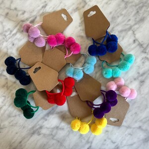May include: Assortment of colourful hair ties with three pom-poms on each tie. The hair ties are displayed on brown card stock with a hole punched at the top.