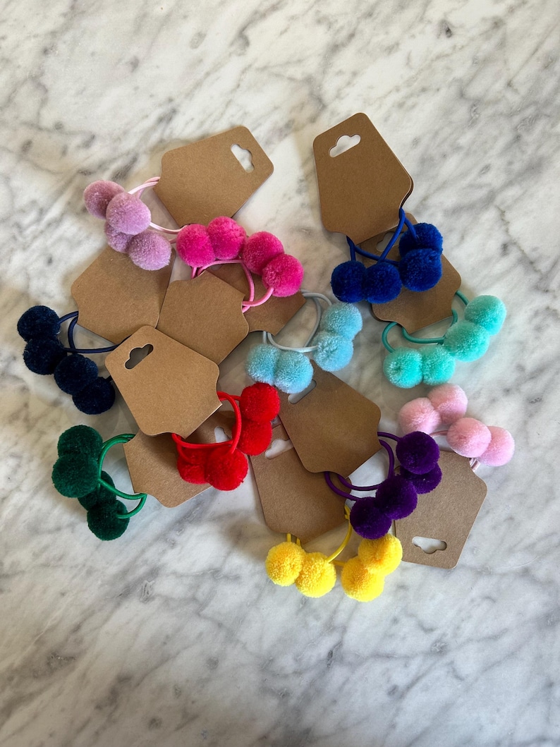May include: Assortment of colourful hair ties with three pom-poms on each tie. The hair ties are displayed on brown card stock with a hole punched at the top.