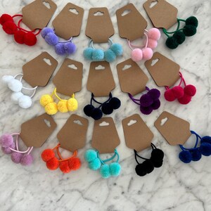 May include: Assortment of colourful pom-pom hair ties on brown card stock. The hair ties are in various colours, including white, purple, pink, green, yellow, orange, blue, and black.