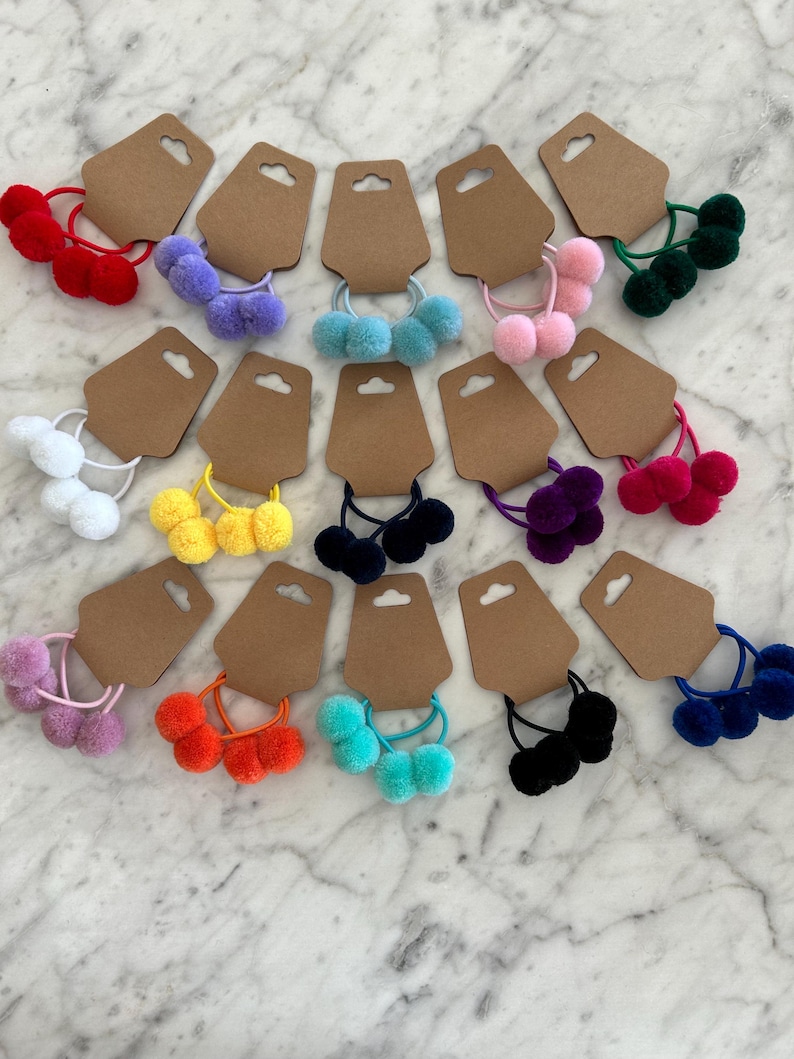 May include: Assortment of colourful pom-pom hair ties on brown card stock. The hair ties are in various colours, including white, purple, pink, green, yellow, orange, blue, and black.