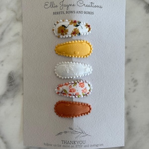 May include: Set of five hair clips with floral patterns. The clips are oval shaped and have a scalloped edge. The patterns include yellow flowers on white, white floral embroidery, pink flowers on white, and solid brown.