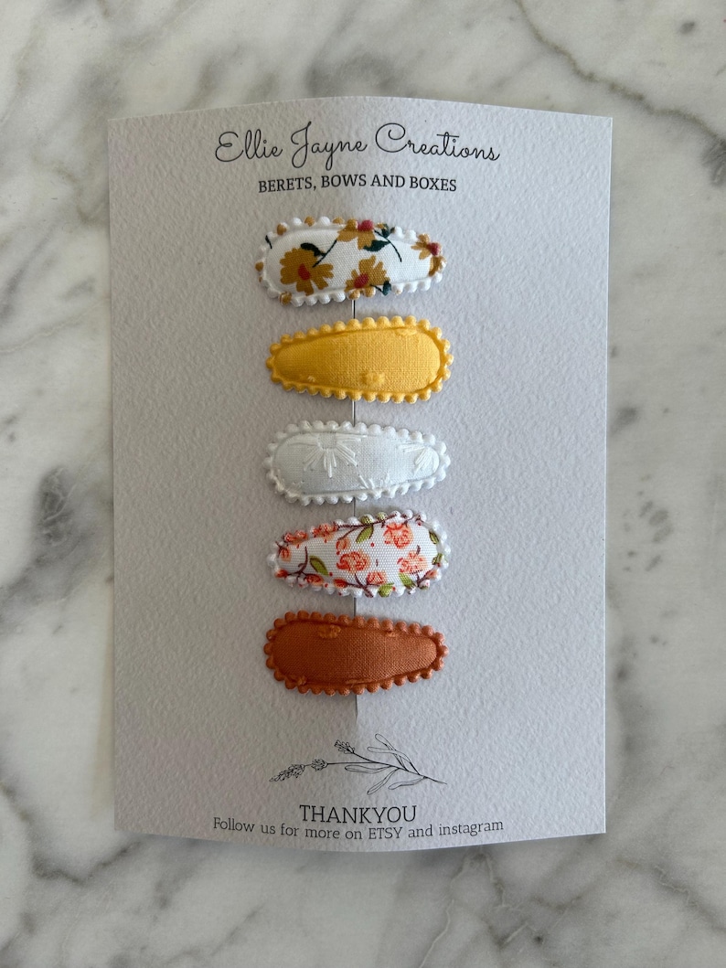 May include: Set of five hair clips with floral patterns. The clips are oval shaped and have a scalloped edge. The patterns include yellow flowers on white, white floral embroidery, pink flowers on white, and solid brown.