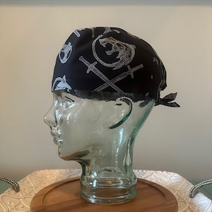 May include: Black scrub hat with a white print of swords and a wolf head. The hat is tied on a mannequin head.