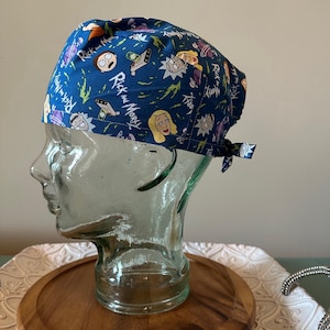 May include: Blue surgical scrub cap with a repeating pattern of cartoon characters and the words "Rick and Morty". The cap is tied at the back and is displayed on a glass head.