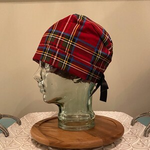 May include: Red, blue, green and yellow plaid surgical scrub hat with black ties. The hat is on a glass head form and a wooden tray.