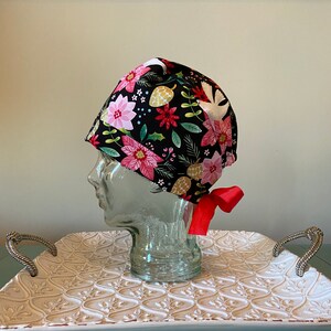 May include: A black scrub hat with a Christmas floral print featuring red poinsettias, green leaves, gold pine cones, and white doves. The hat has a red ribbon tie.