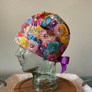 May include: A colourful surgical scrub hat with a donut pattern. The hat is tied with a purple ribbon.