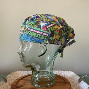 Peut inclure: Un bonnet de chirurgien bleu et vert avec un imprimé de bande dessinée Teenage Mutant Ninja Turtles. Le bonnet a une cravate à l'arrière.