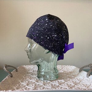 May include: A black scrub hat with a white and purple splatter pattern. The hat has a purple ribbon tied at the back.