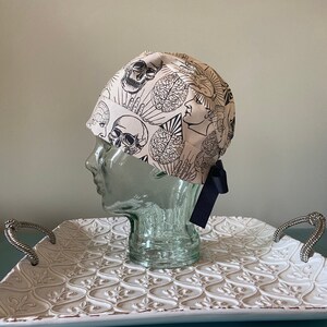 May include: A beige surgical scrub cap with a black and white anatomical design of skulls, brains, and profiles. The cap has a dark blue ribbon tie at the back. The cap is displayed on a glass head form.