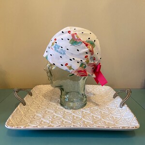 May include: White surgical scrub hat with a black polka dot pattern and a floral print. The hat has a pink bow tied on the side.