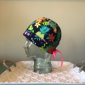 May include: A blue surgical scrub hat with a tropical print featuring pineapples, flowers, and leaves. The hat has a pink tie.