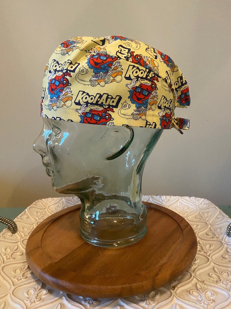 May include: Yellow surgical scrub hat with a repeating pattern of Kool-Aid pitchers wearing sunglasses and riding skateboards. The Kool-Aid logo is printed on the hat.