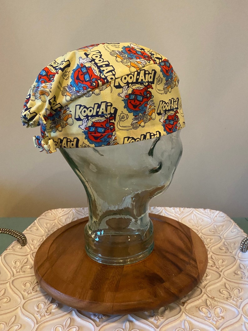 May include: Yellow surgical scrub hat with a repeating pattern of Kool-Aid Man cartoon characters and the Kool-Aid logo.