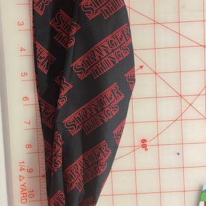 May include: Black fabric with a red "Stranger Things" pattern. The fabric is folded and laid on a white cutting mat with red grid lines.