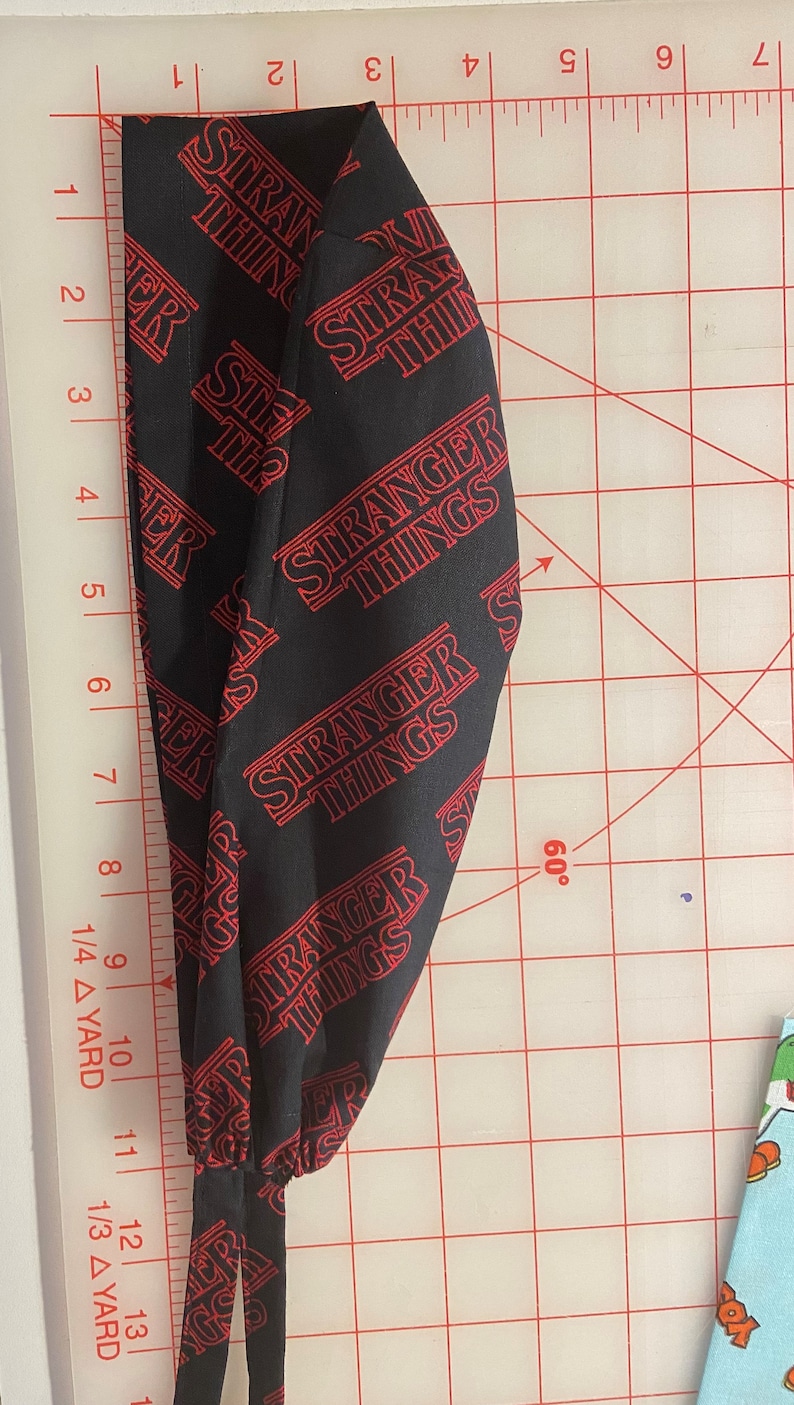May include: Black fabric with a red "Stranger Things" pattern. The fabric is folded and laid on a white cutting mat with red grid lines.