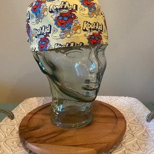 May include: A yellow scrub hat with a repeating pattern of Kool-Aid characters and the Kool-Aid logo. The hat is on a glass head form.