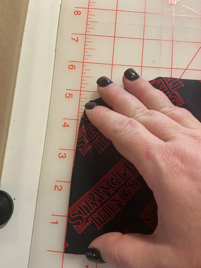 May include: Black fabric with the words "Stranger Things" repeated in red. The fabric is being folded by a hand with black nail polish.