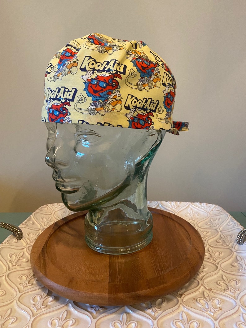 May include: A yellow surgical scrub hat with a repeating pattern of the Kool-Aid mascot and the Kool-Aid logo.