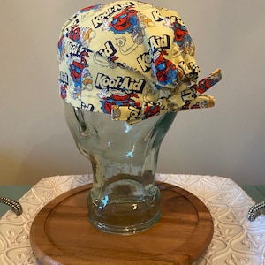 May include: A yellow surgical scrub hat with a repeating pattern of the Kool-Aid logo and a cartoon character. The hat is tied in a bow at the back.
