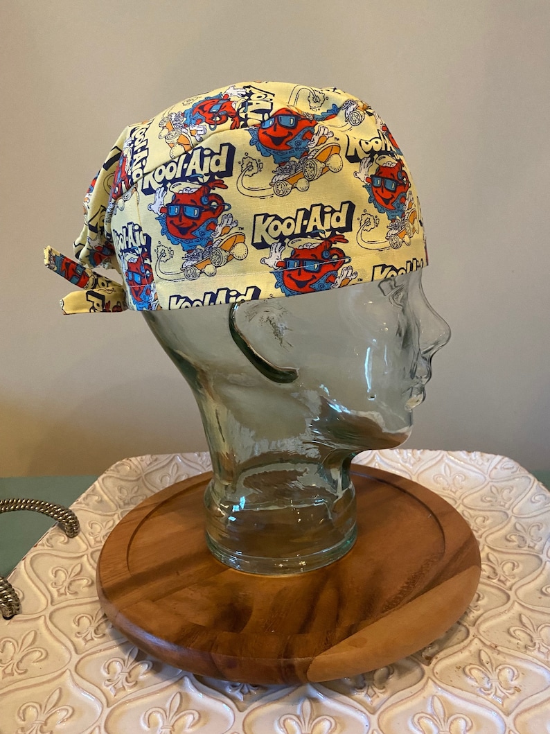 May include: Yellow fabric scrub hat with a repeating pattern of Kool-Aid smiling pitcher characters wearing sunglasses and riding skateboards.