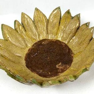 May include: A ceramic bowl shaped like a sunflower with a dark brown textured center and yellow petals radiating outward. The petals feature a glossy finish with subtle green highlights along the edges, creating a realistic floral appearance.